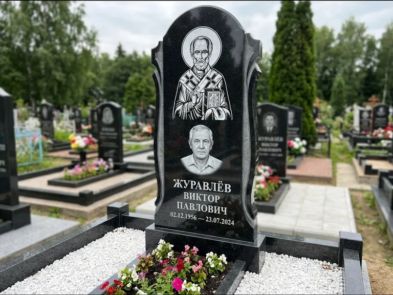 Памятник с гравировкой иконы: стела с образом в полный рост на обороте — гравировка образа Матроны Московской, низкие кусты спиреи в цвету, работа «Лебедева» в Нижневартовске