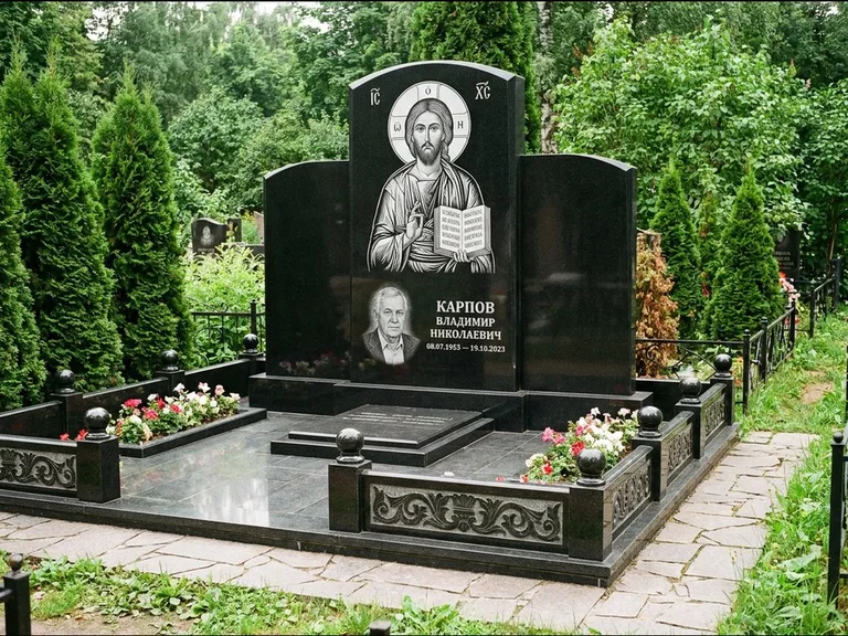 Памятник с гравировкой иконы: широкая панорамная стела с золочёным нимбом — гравировка образа Матроны Московской, цветник с белыми хризантемами, работа «Мельникова» в Нижневартовске