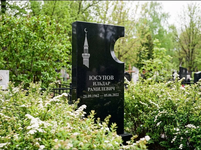 Мусульманский памятник с гравировкой мечети: стела с боковым вырезом в форме полумесяца — гравировка минарета с полумесяцем, низкие кусты спиреи в цвету, работа «Юсупов» в Нижневартовске